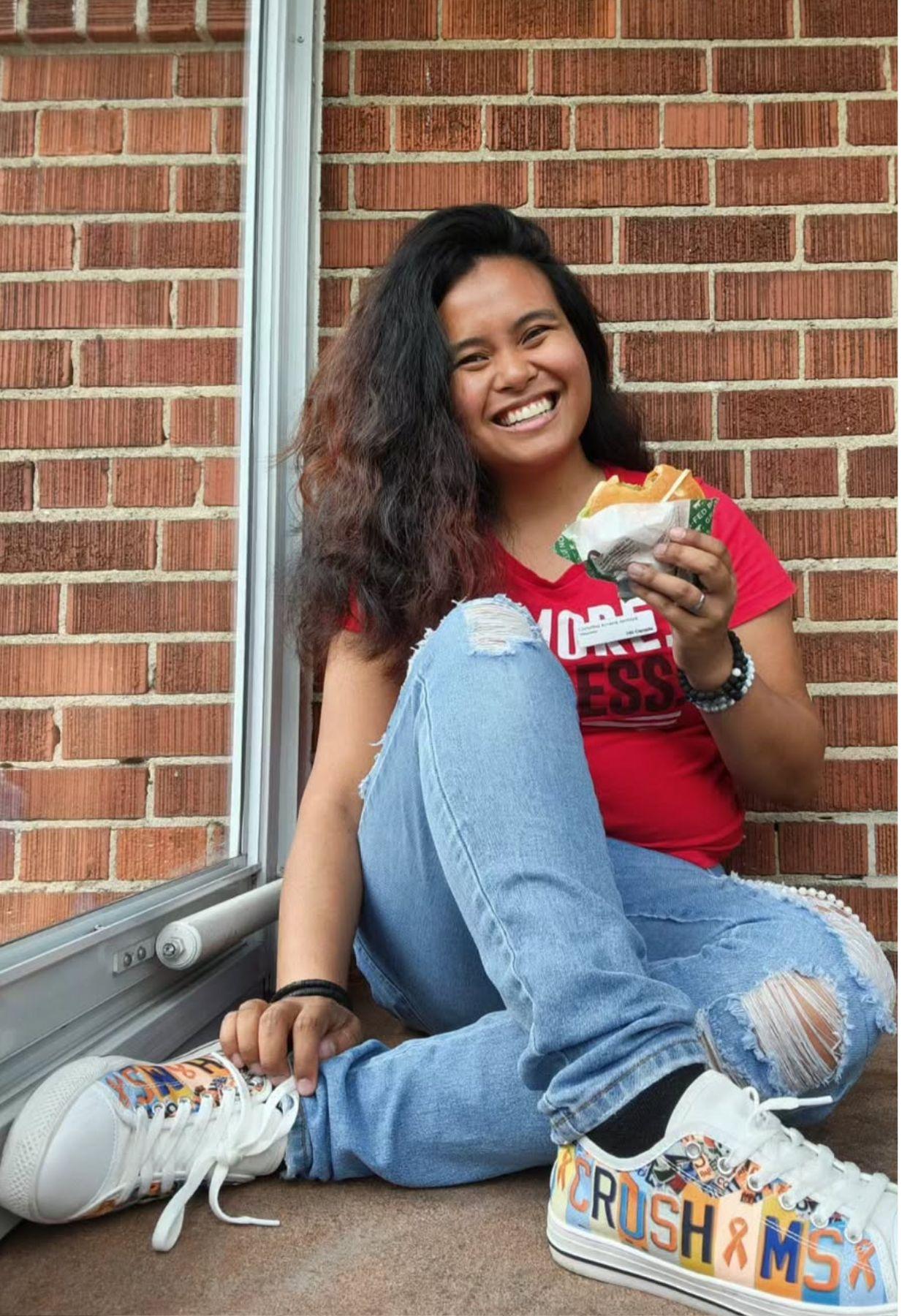 Christina sitting on the ground, wearing her "Crush MS" shoes, smiling and holding a Teen Burger®