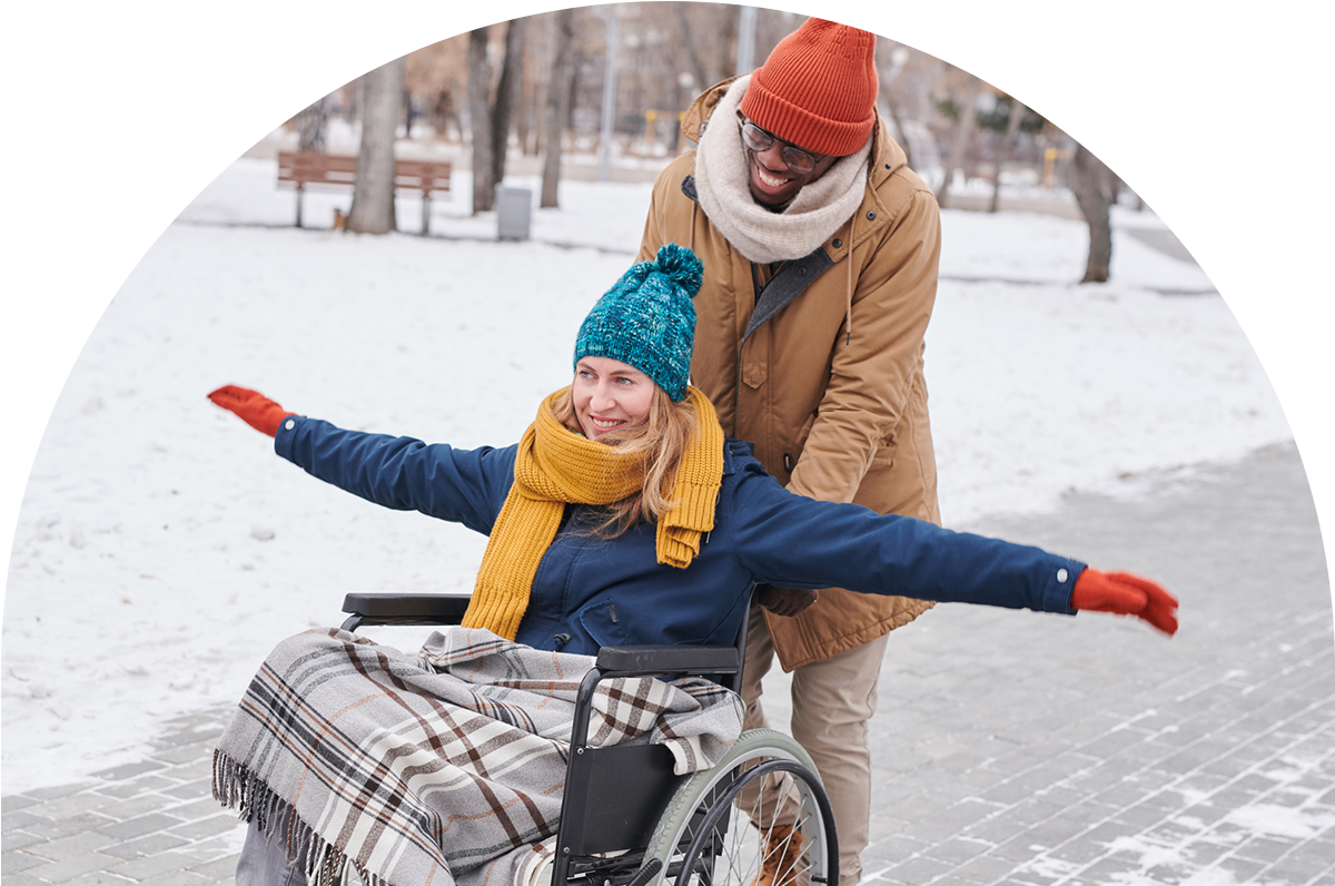 A man in a brown coat, scarft and orange toque pushes a woman in a wheelchair who is wearing a blue coat with a teal toque, red mittens and a yellow scarf. It is snowy.