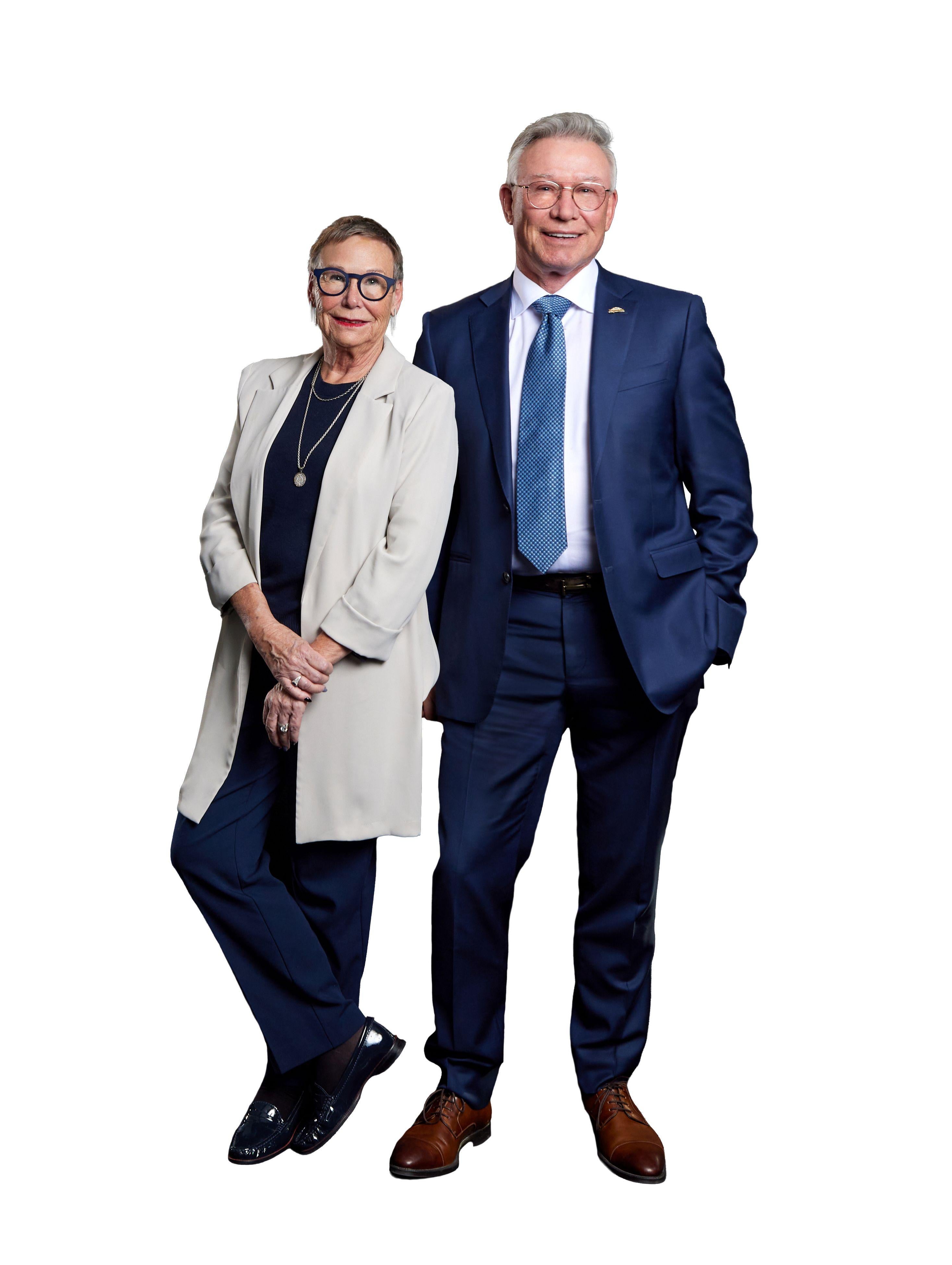 Diana wearing glasses, long cream blazer, dark blue shirt and pants, standing beside Jay who's wearing glasses, a dark blue suit with a white shirt and blue tie