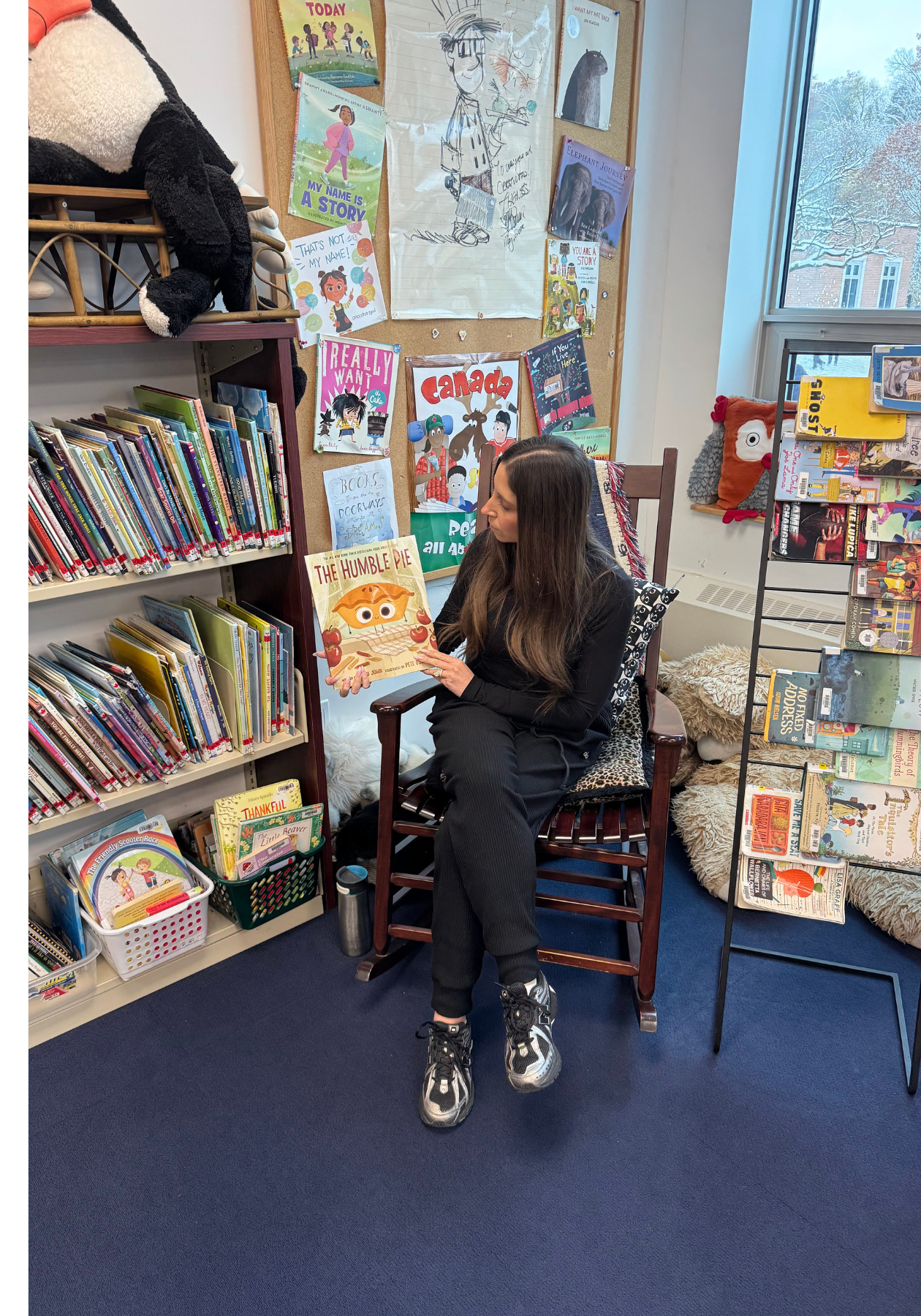 Jaclyn, wearing a black top and black pants, sitting in a chair, holding up a book.