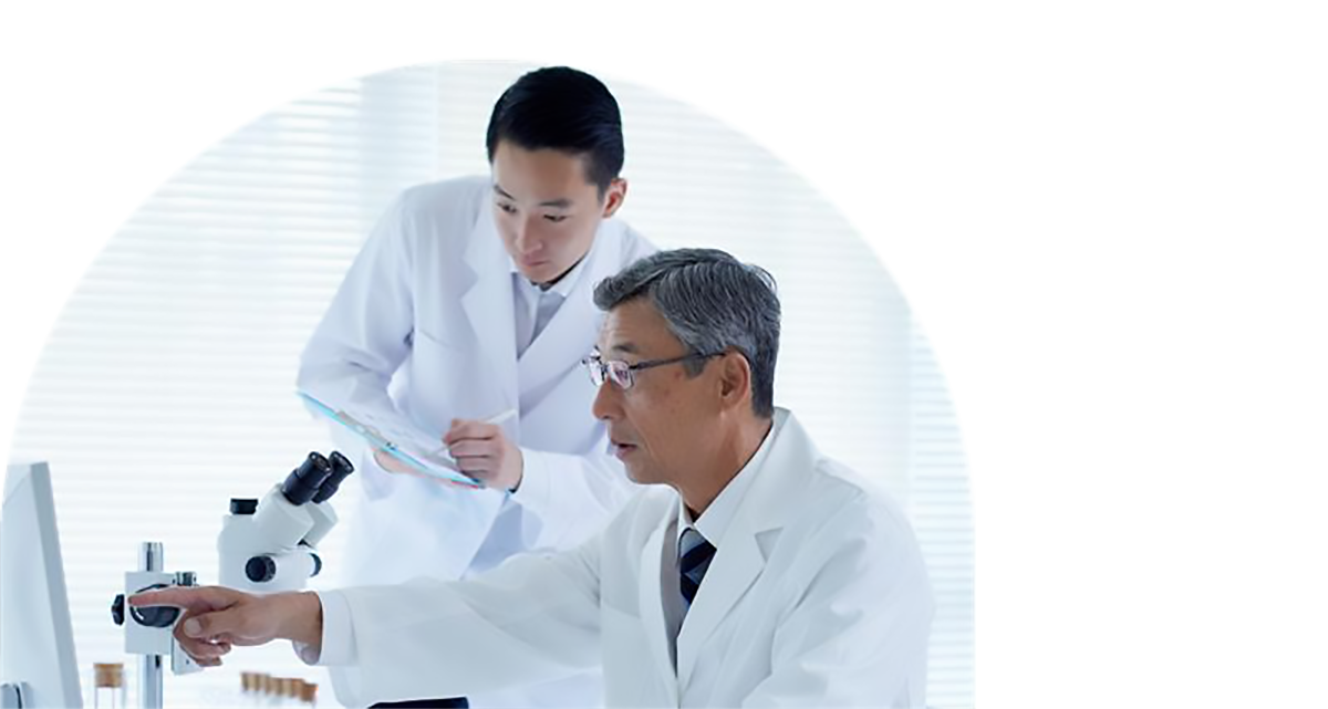 Two researchers in white lab coats. One wearing glasses and pointing at a computer screen and one holding a clipboard and pen.