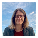 Professional headshot of a woman with glasses and shoulder-length blonde hair against a cloudy sky.