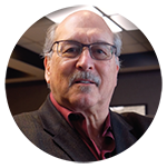 Circular portrait of an older man with glasses wearing a dark blazer.
