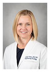 Professional headshot of a smiling blonde woman in a white medical coat.