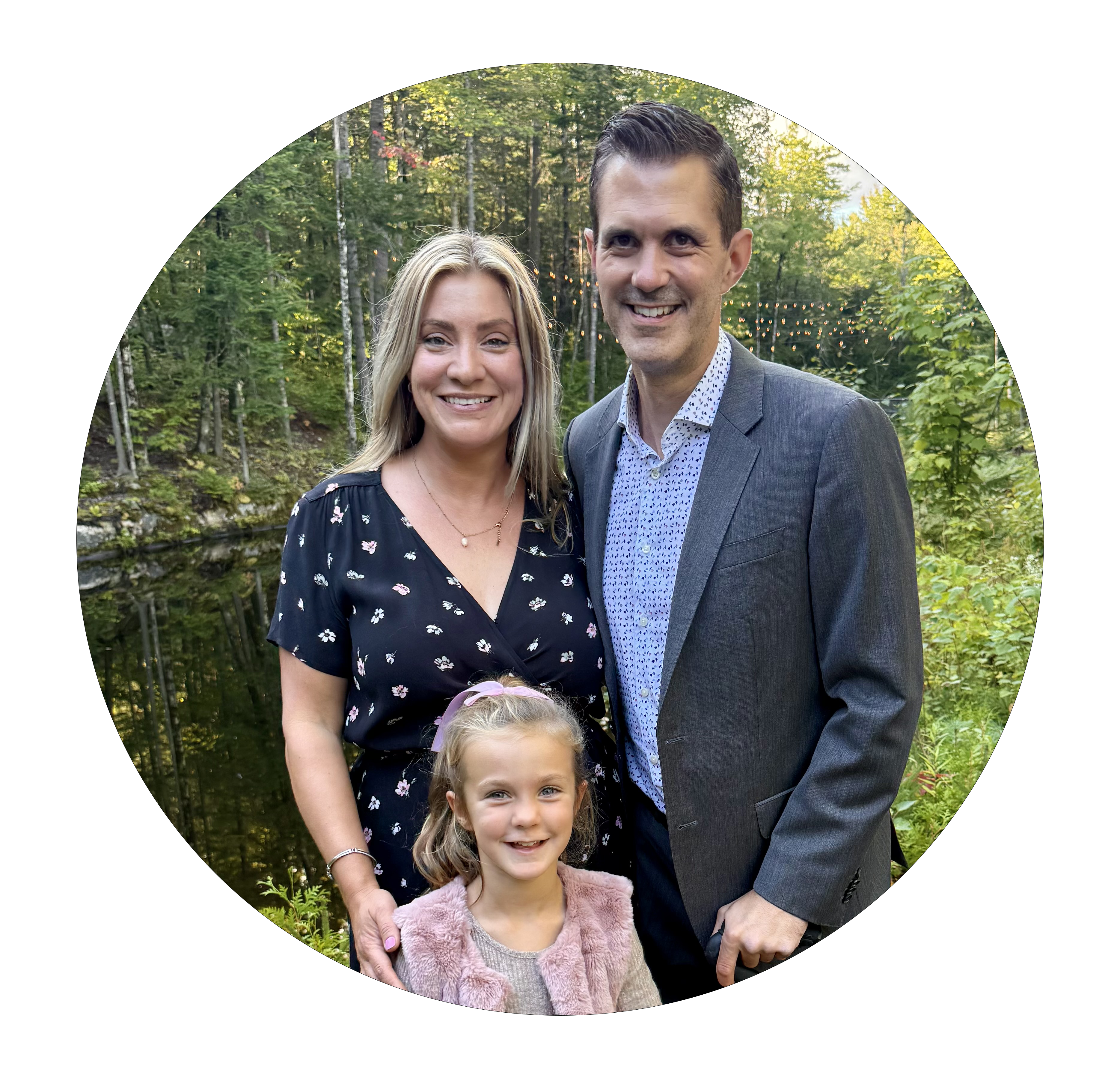 Smiling family of three outdoors by a wooded pond, parents with young blonde girl.