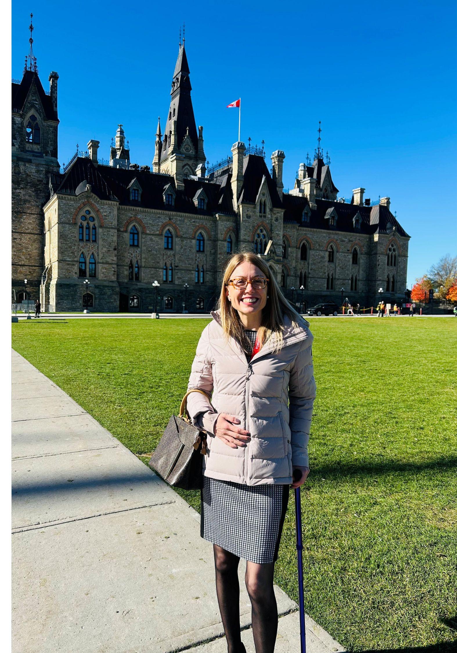 Candice smiling in front of Parliament Hill on a sunny day, wearing glasses, a beige jacket, checkered skirt, and holding a cane.