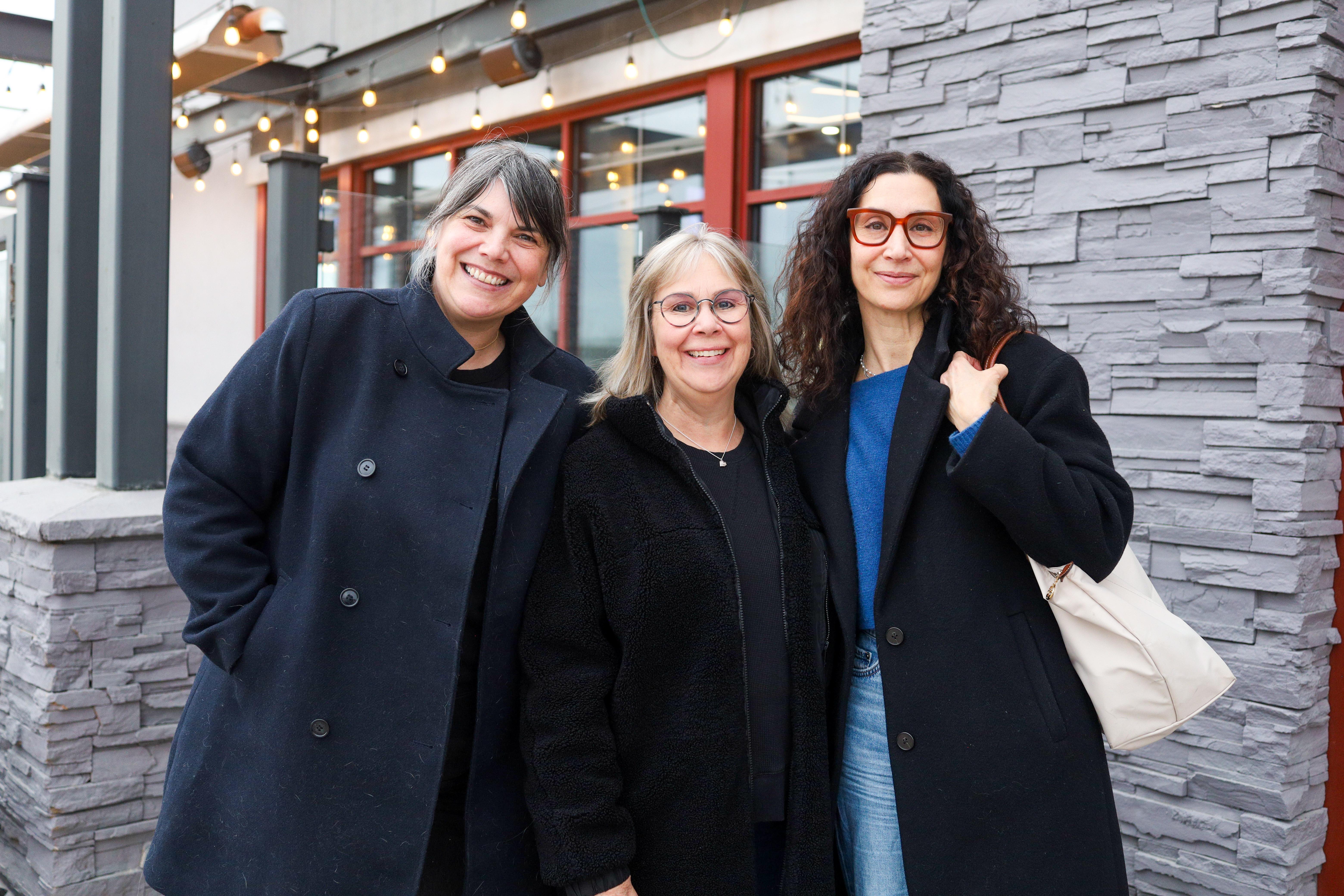Three women smiling together outdoors, wearing dark coats on a cloudy day.
