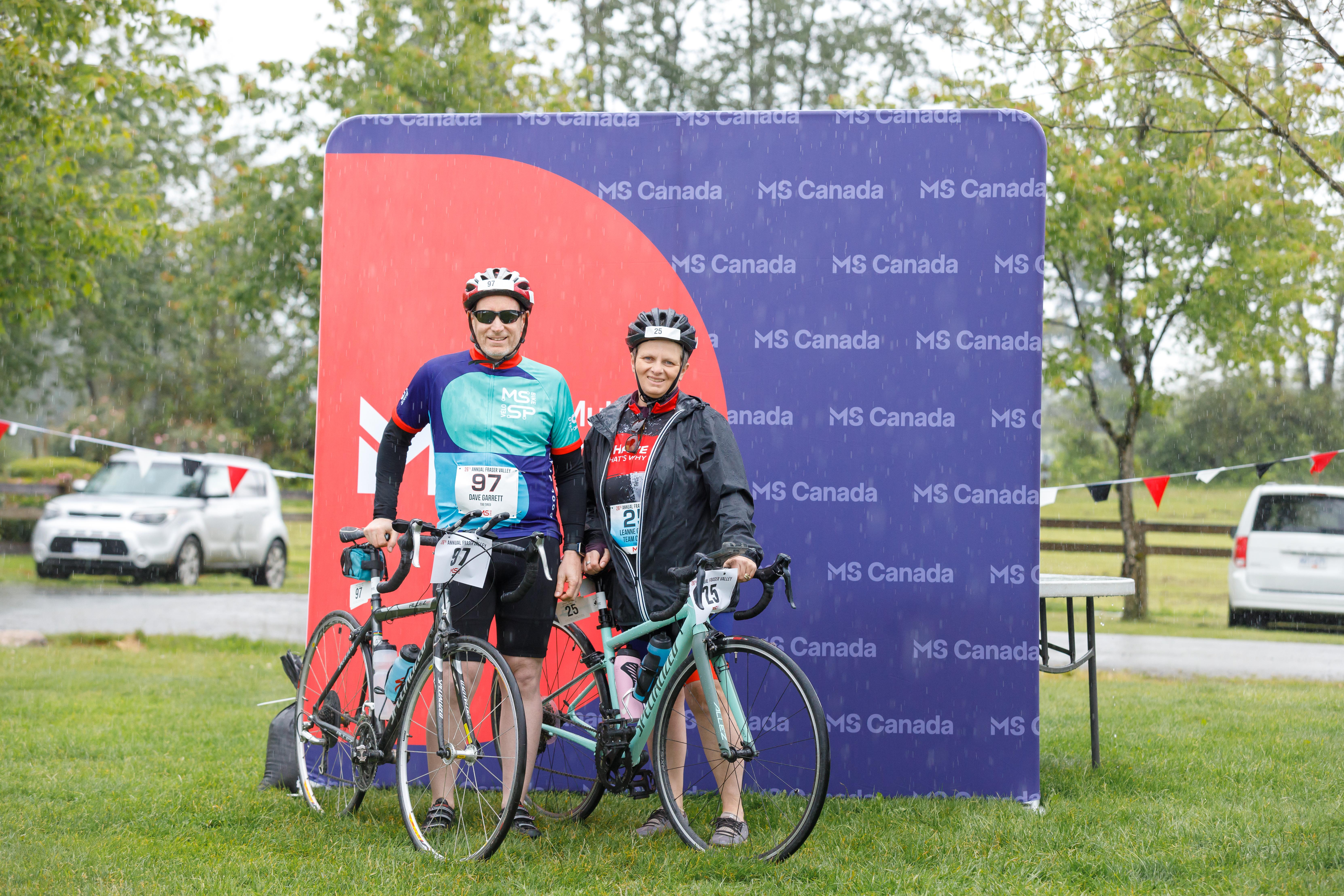 Leanne and her husband wearing MS Bike jerseys with road bikes posing in front of the MS Canada banner.