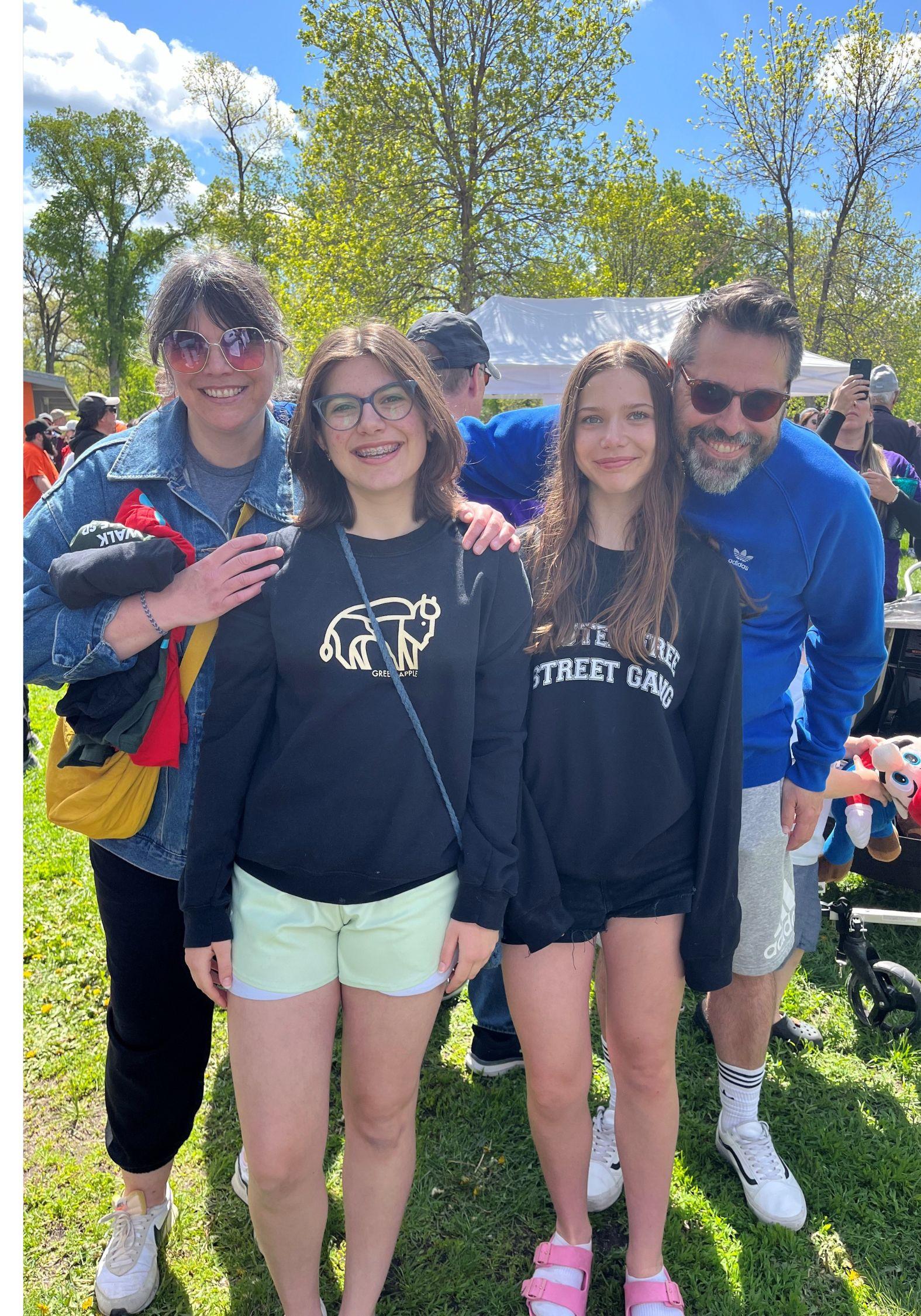 Four smiling people posing together outdoors on a sunny day with green trees in the background.