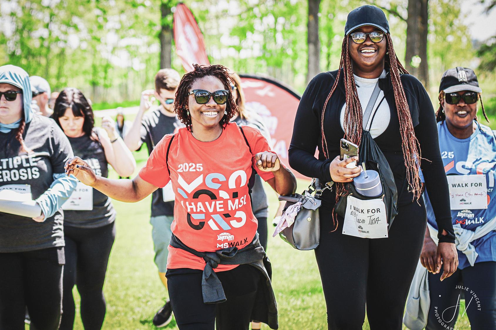 Two women smiling and walking together at an outdoor charity run event on a sunny day.