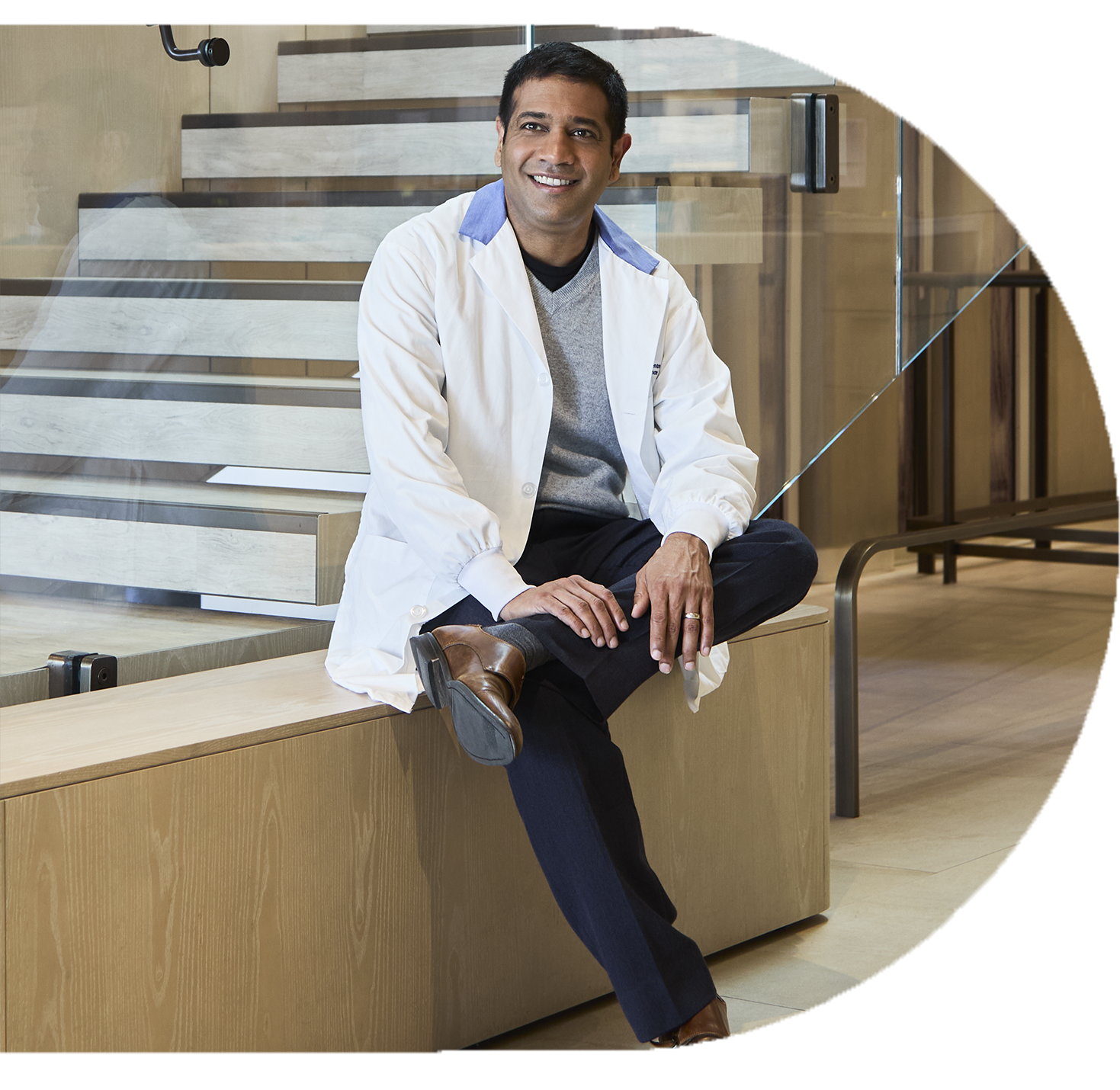 Smiling man in a white lab coat sitting casually on a bench near a staircase.