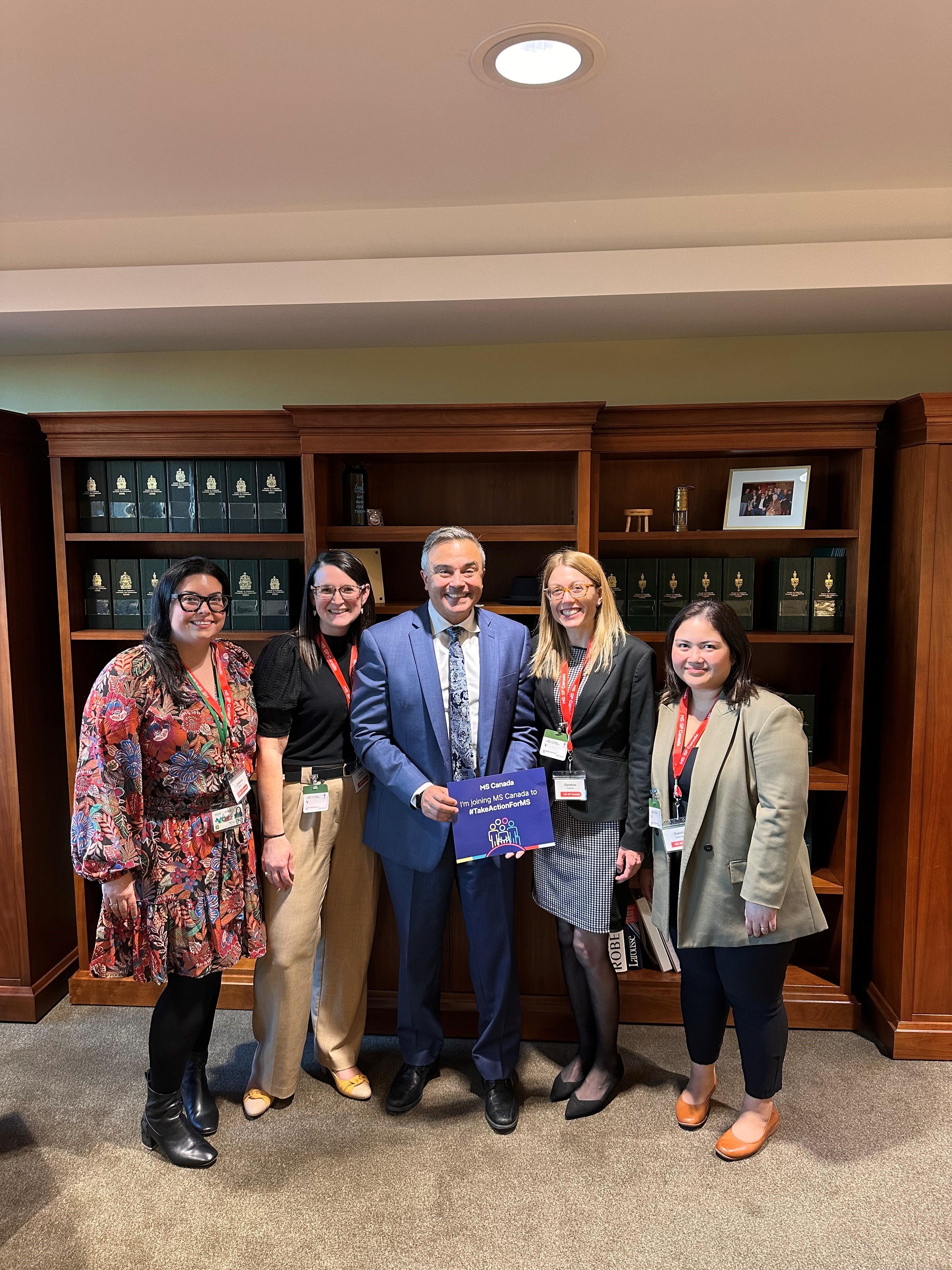Five smiling professionals posing together in an office with wooden bookshelves in the background.