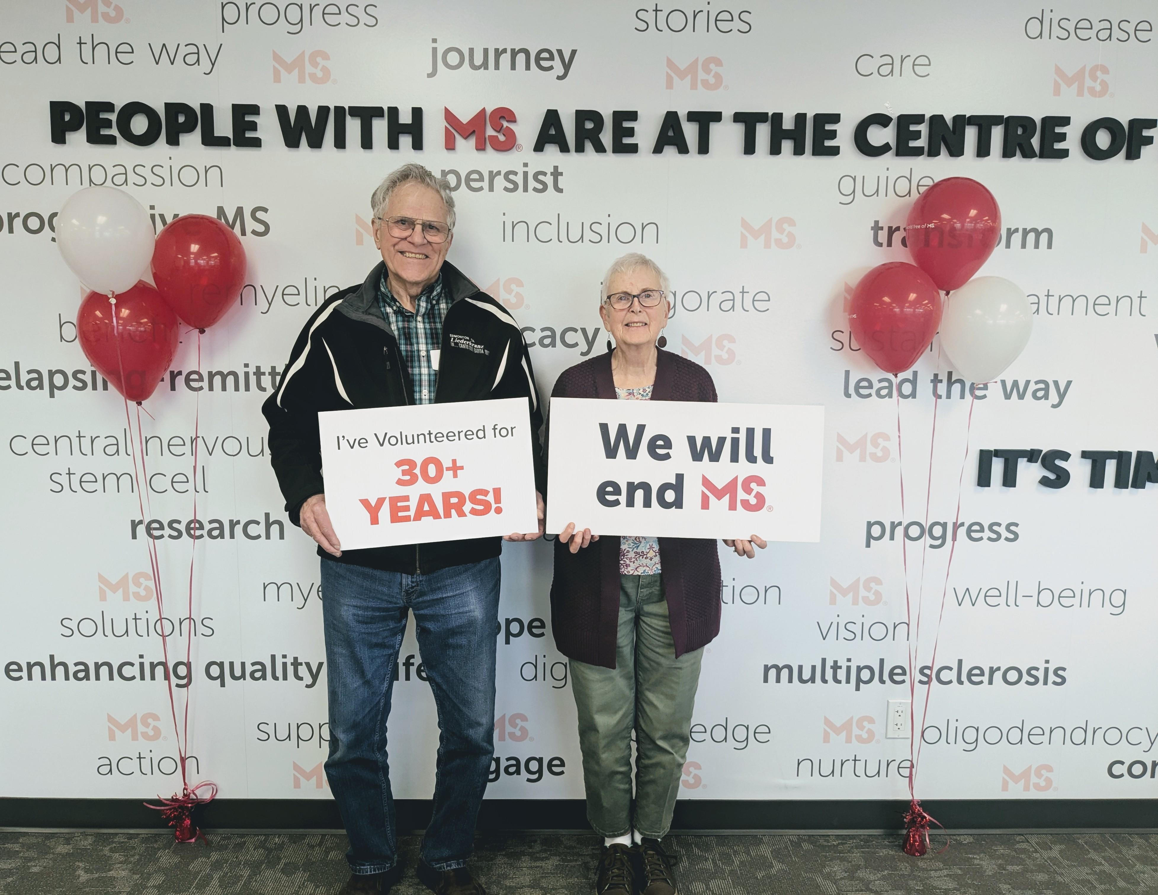 Len holding a sign that says, "I've volunteered for 30+ years" and Sharon holding a sign that says, "We will end MS" 
