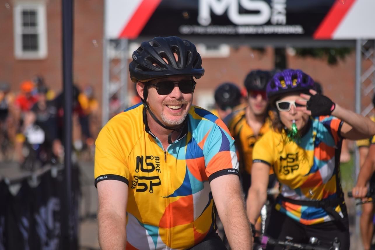 Craig smiling on his bike at the start line, wearing an MS Bike jersey, black helmet, and sunglasses.