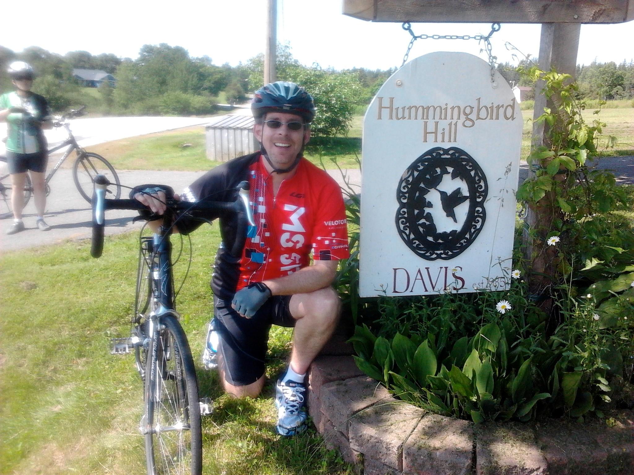 Craig posing with his bike next to a "Hummingbird Hill" sign outdoors.