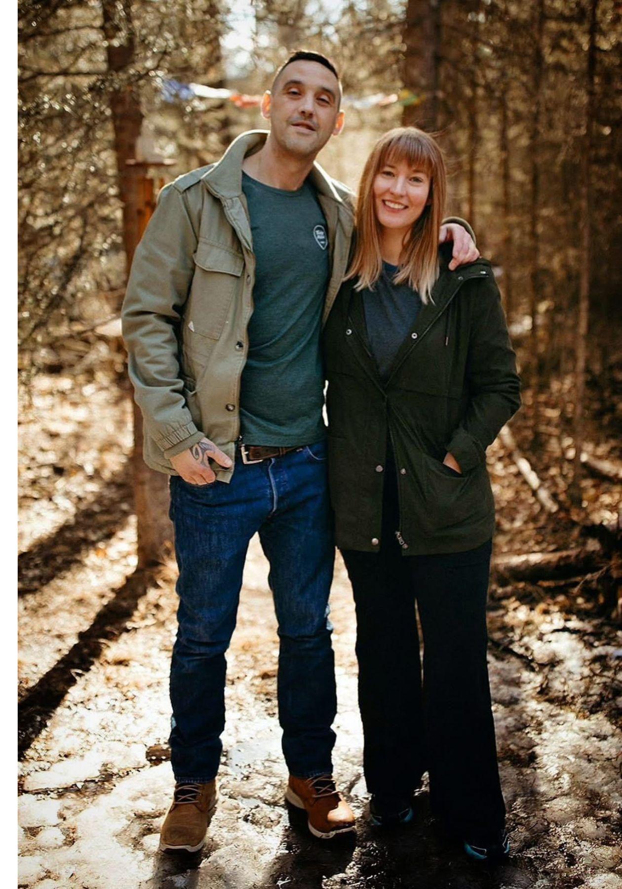 Smiling couple standing together in a snowy forest, wearing casual winter jackets.