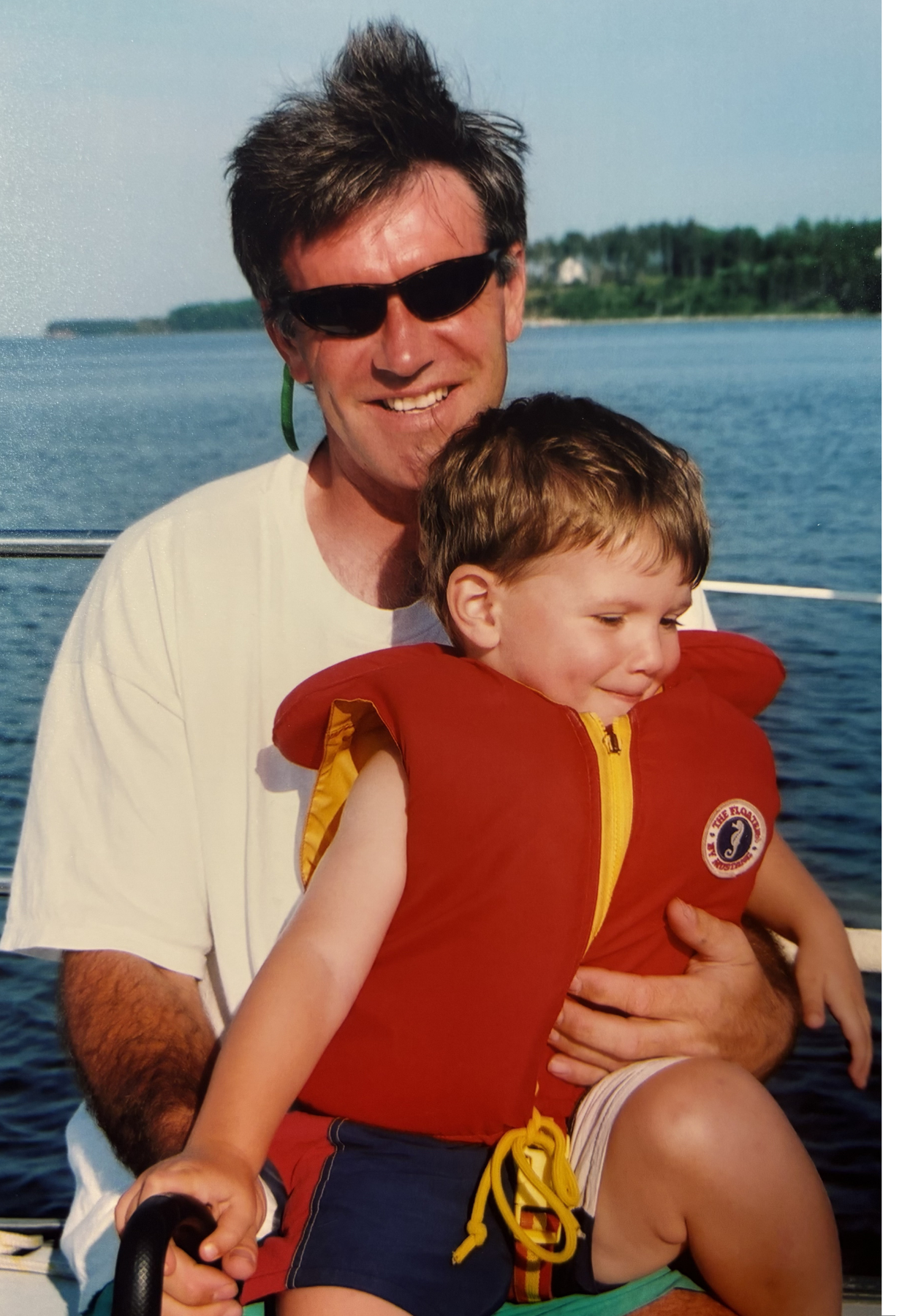 Larry in sunglasses holding his son Glen in a red life jacket on a boat.
