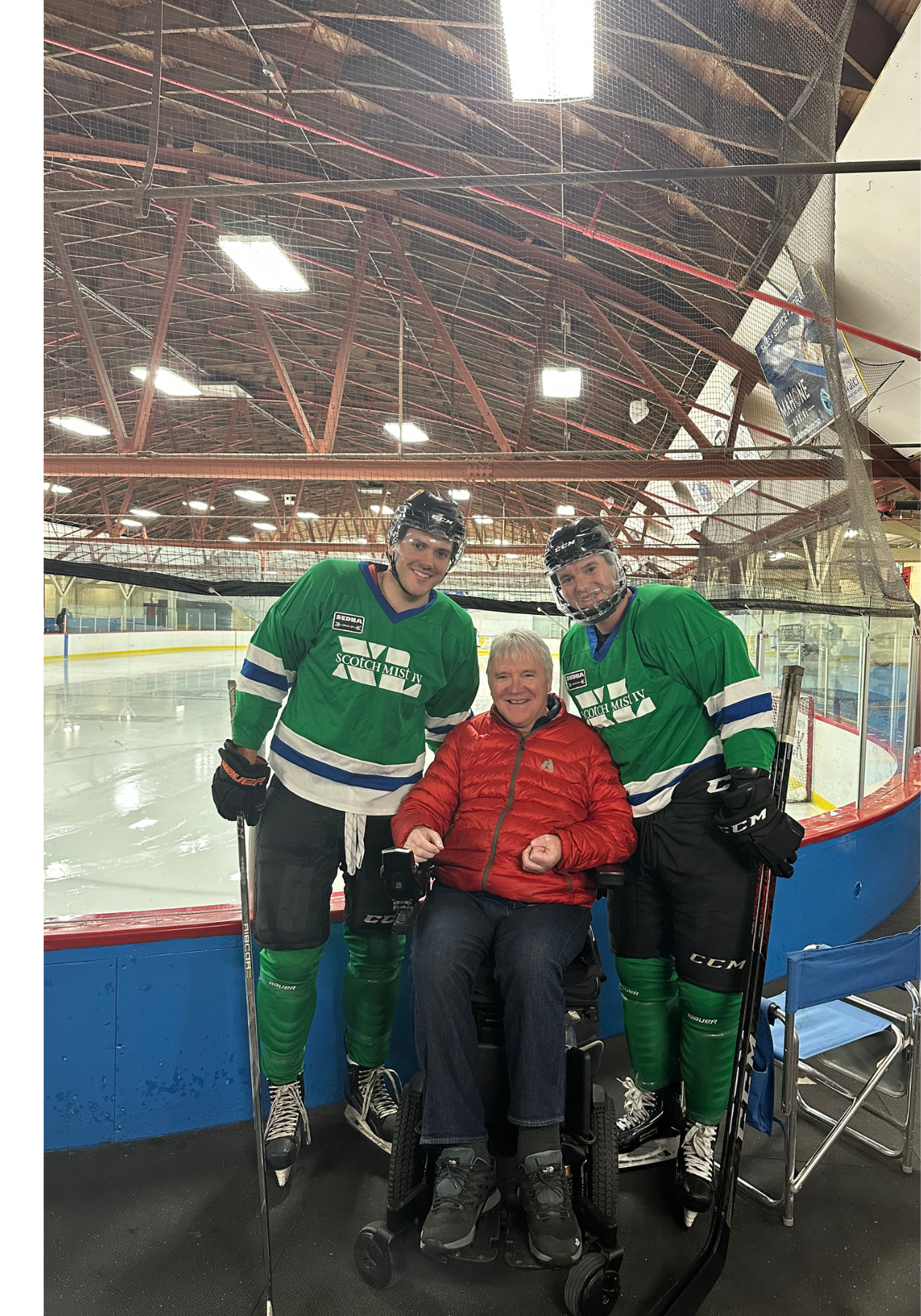 Larry in red jacket seated between his sons in green jerseys at an indoor rink.