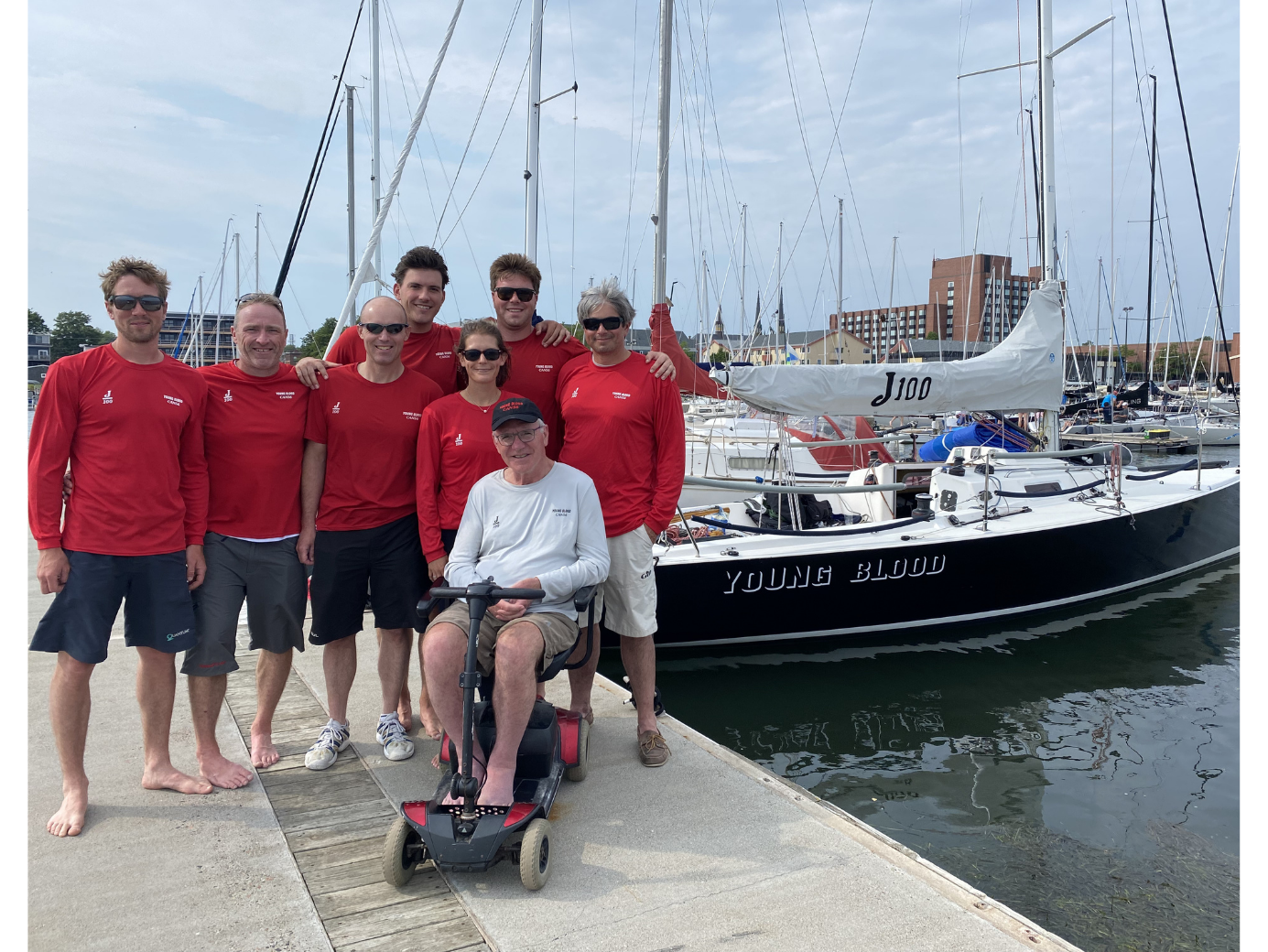 Larry, in a wheelchair, and sailing crew in red shirts posing beside a sailboat at a marina.