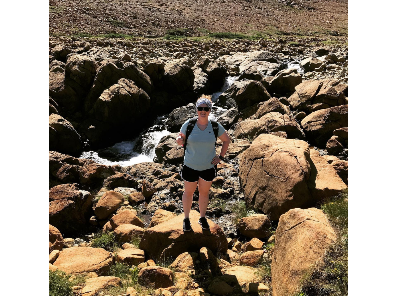 Janice standing on rocky terrain beside a small mountain waterfall on a sunny day.