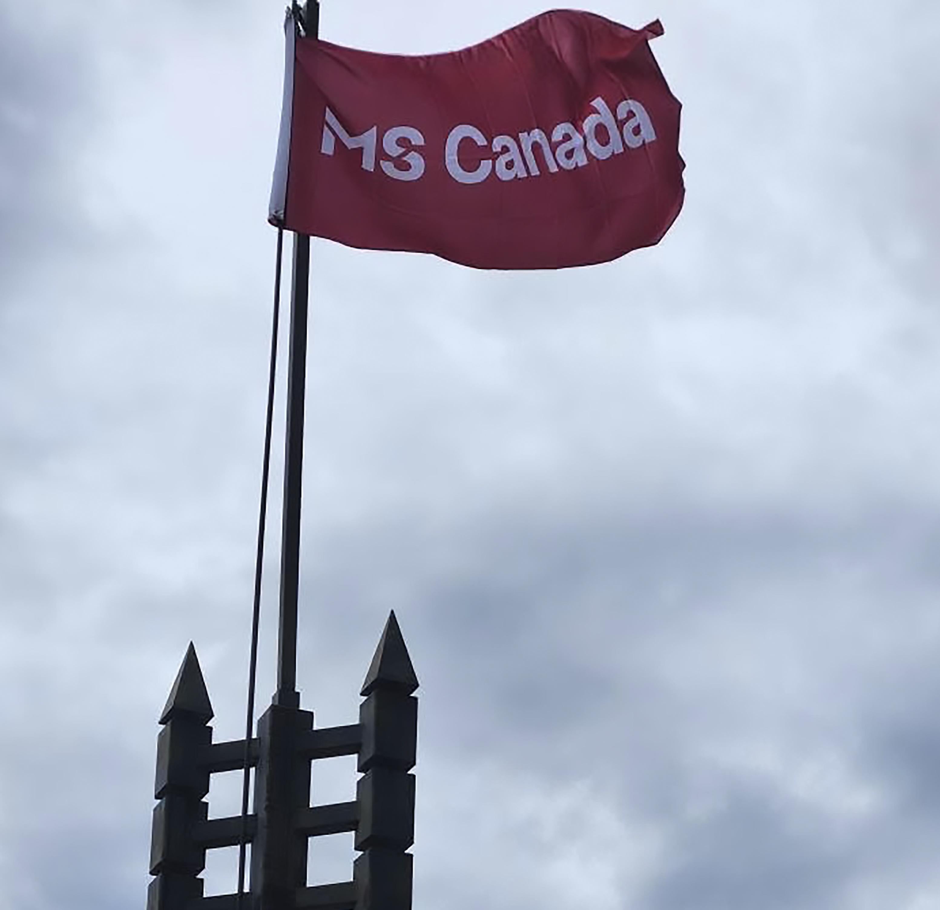 Red MS Canada flag waving on a flagpole against a cloudy sky.