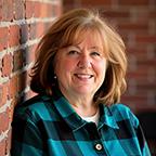 Smiling middle-aged woman in a teal plaid top, posed against a red brick wall.