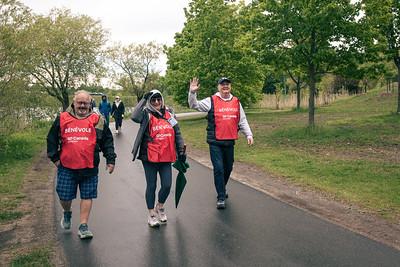 Three people walking on a path in a park, wearing red volunteer vests, surrounded by green trees.