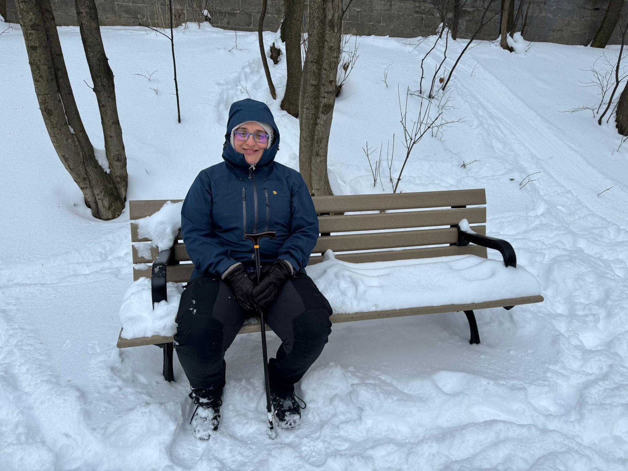 Ksenia in winter gear sitting on a snow-covered park bench outside.
