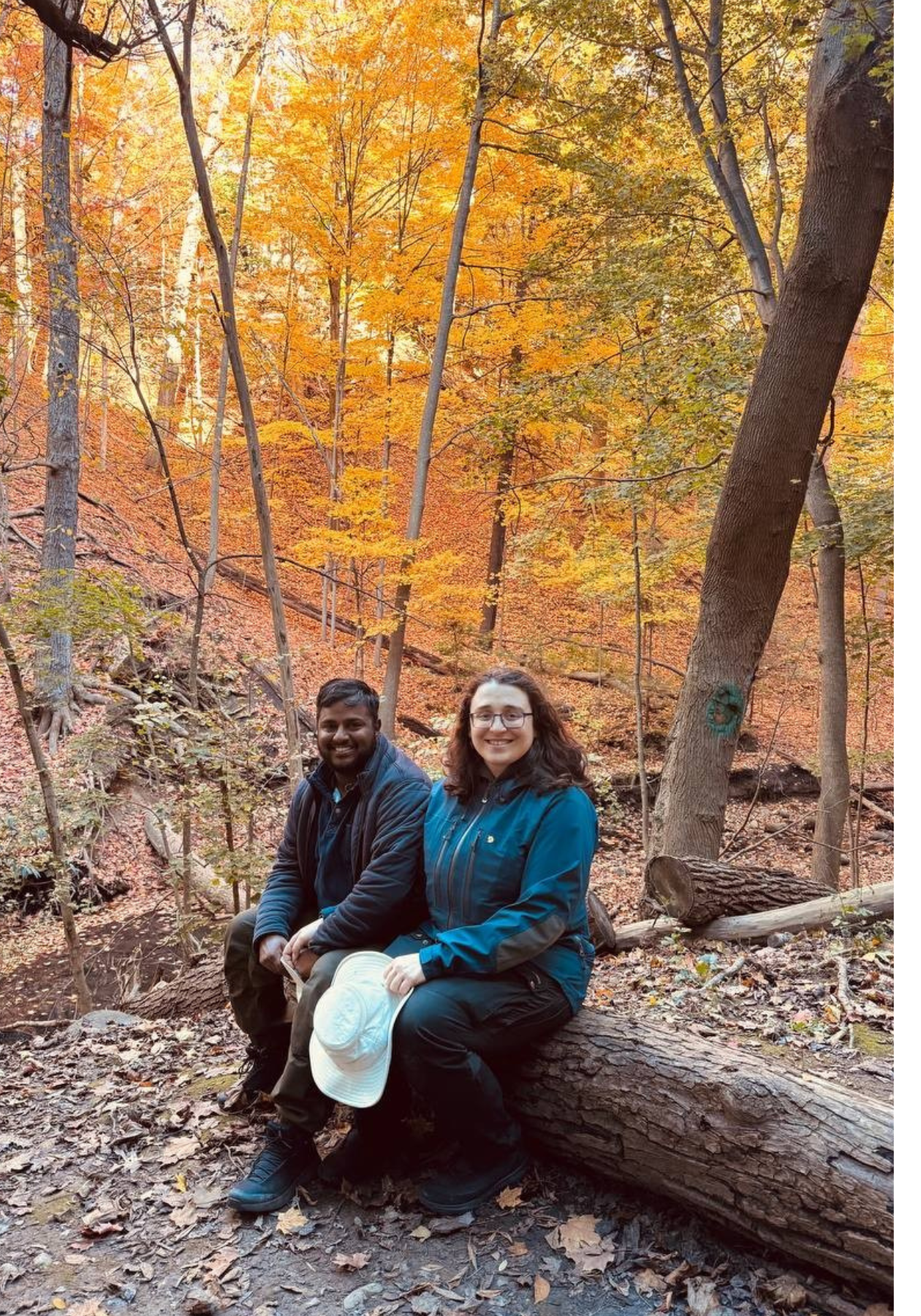 Ksenia (right) and her partner Megh sit on a tree log outside in front of autumn coloured trees.