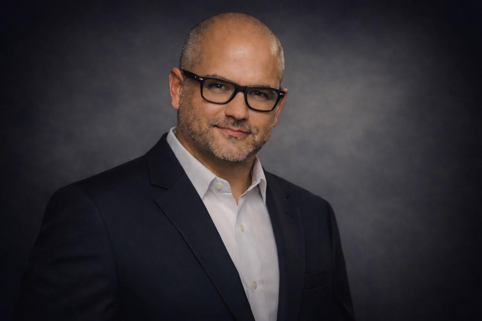 Professional headshot of Dario in glasses and a dark blazer against a gray background.