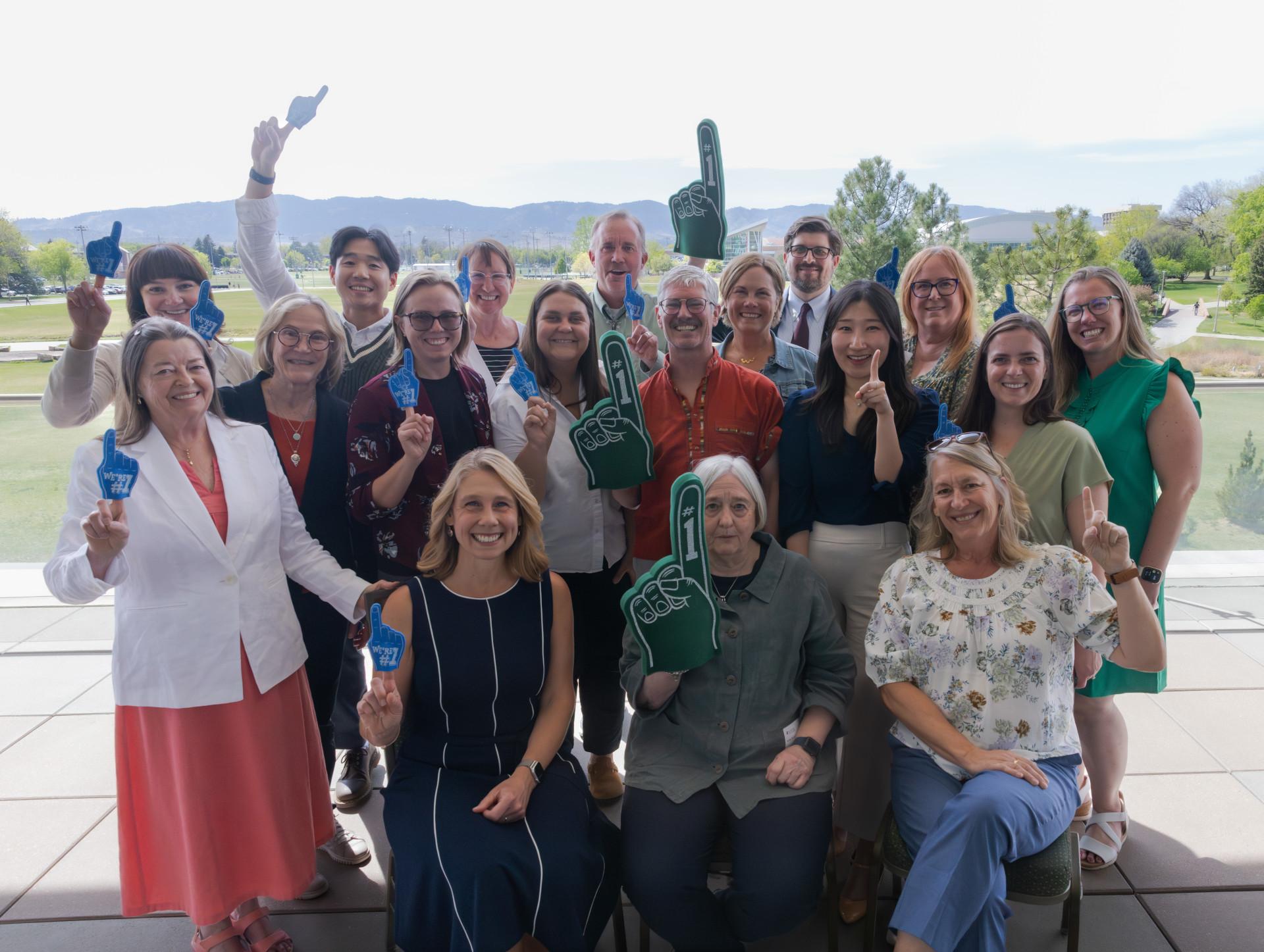 Paul and his colleagues at the Department of Occupational Therapy at Colorado State University are celebrating and wearing green and blue foam fingers. The group is standing outside in front of a park with grass and trees, and it’s sunny.