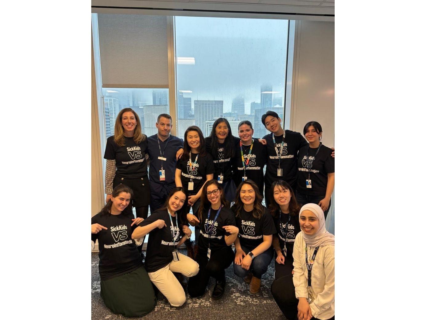 Paul during his Postdoctoral Fellowship at the SickKids Pediatric Neuroinflammatory Disorders Program. Paul and his colleagues are smiling and wearing black and white shirts that read SickKids vs Neuroinflammation. The group is standing on carpet, in front of a large window overlooking tall city buildings.