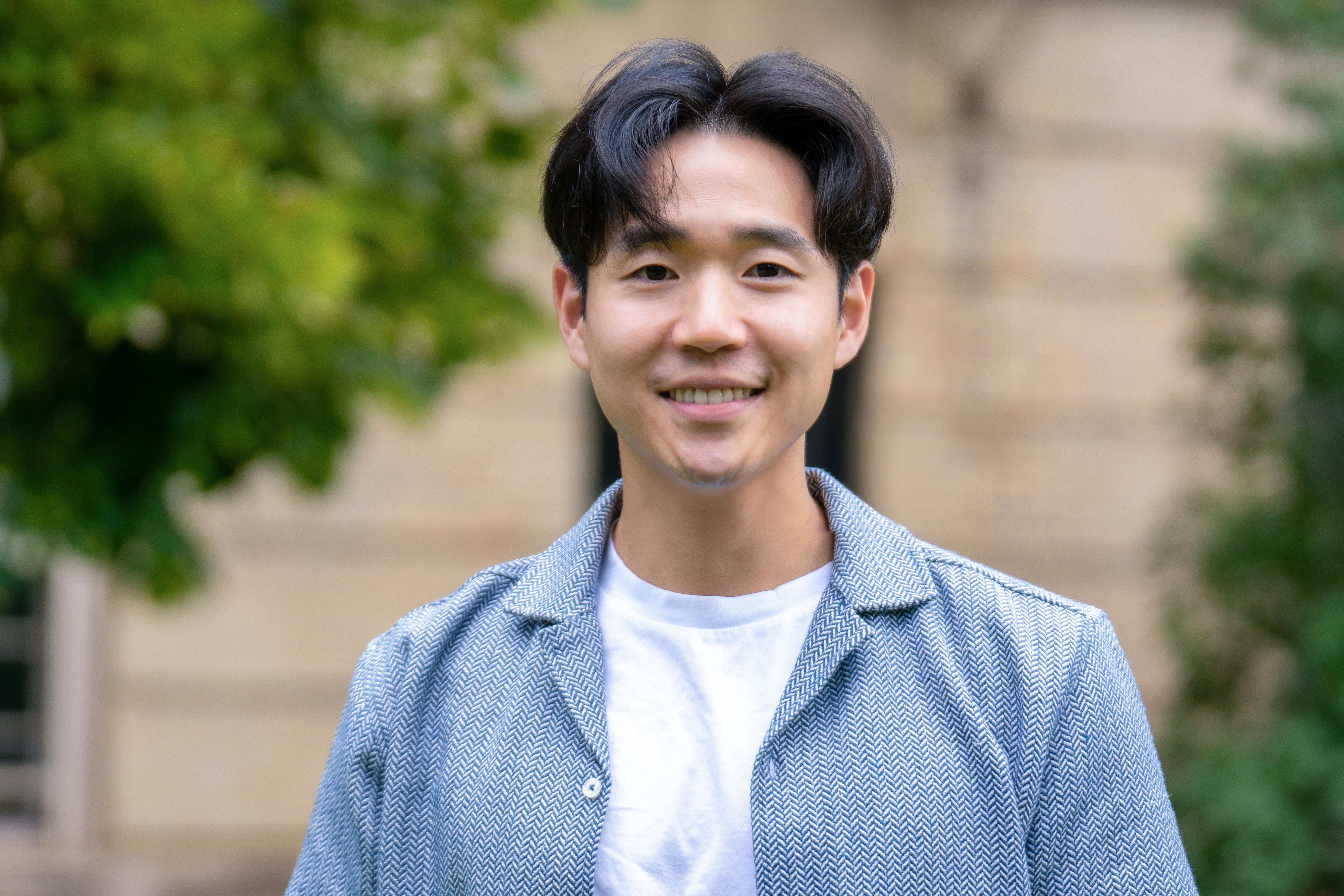 Dr. Paul Yoo is wearing a white t-shirt with a light blue blazer. He's smiling and standing in front of a building and trees.