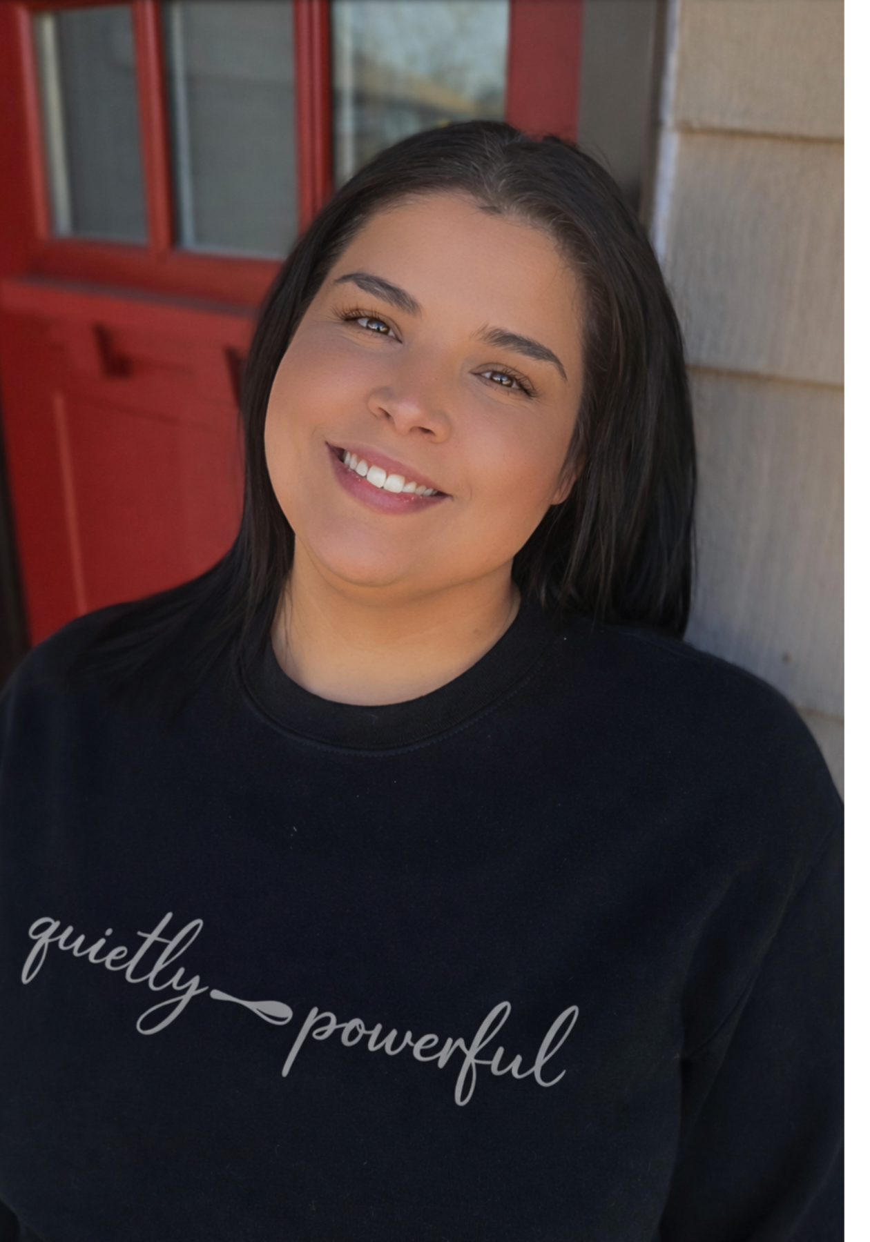 Rachel smiling in a black "quietly powerful" sweatshirt, standing near a red door.