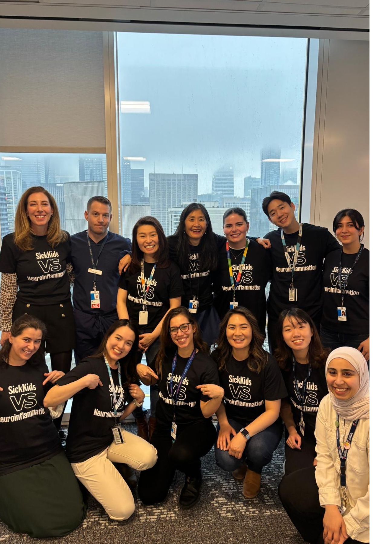 Paul during his Postdoctoral Fellowship at the SickKids Pediatric Neuroinflammatory Disorders Program. Paul and his colleagues are smiling and wearing black and white shirts that read SickKids vs Neuroinflammation. The group is standing on carpet, in front of a large window overlooking tall city buildings.