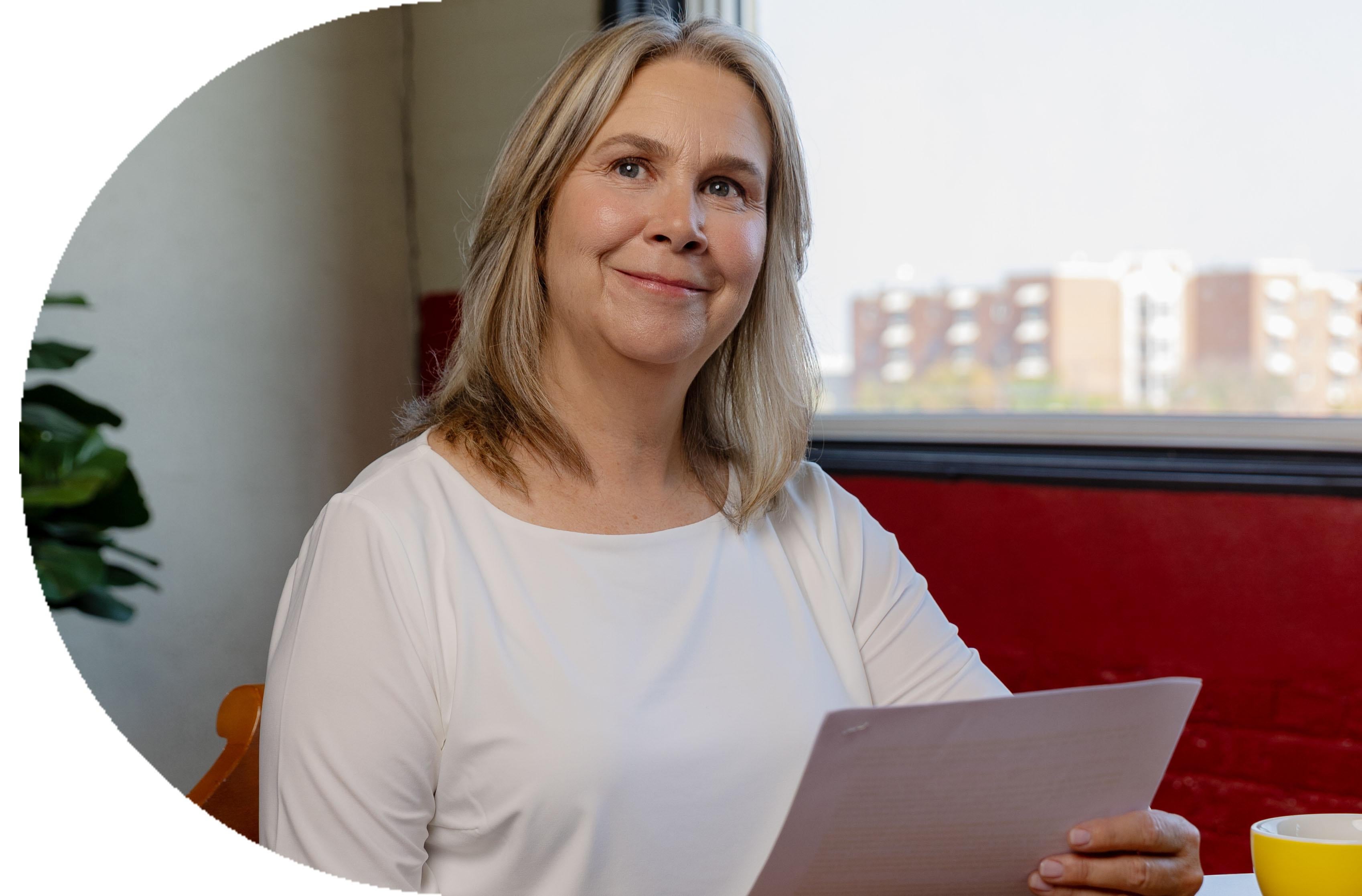 Woman with blonde hair holding documents, seated near a bright window indoors.