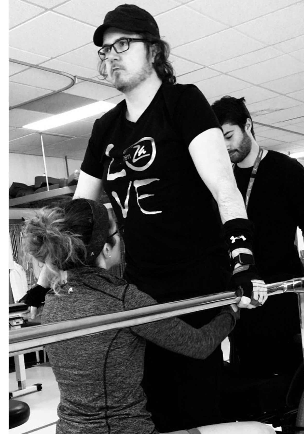 Black and white photo of Jason in a "Love" shirt using a balance bar exercise at physical therapy.