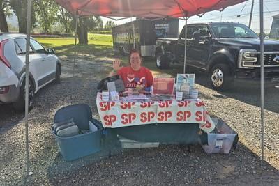Person sitting at an outdoor vendor table displaying "SP" branded products under a red canopy.