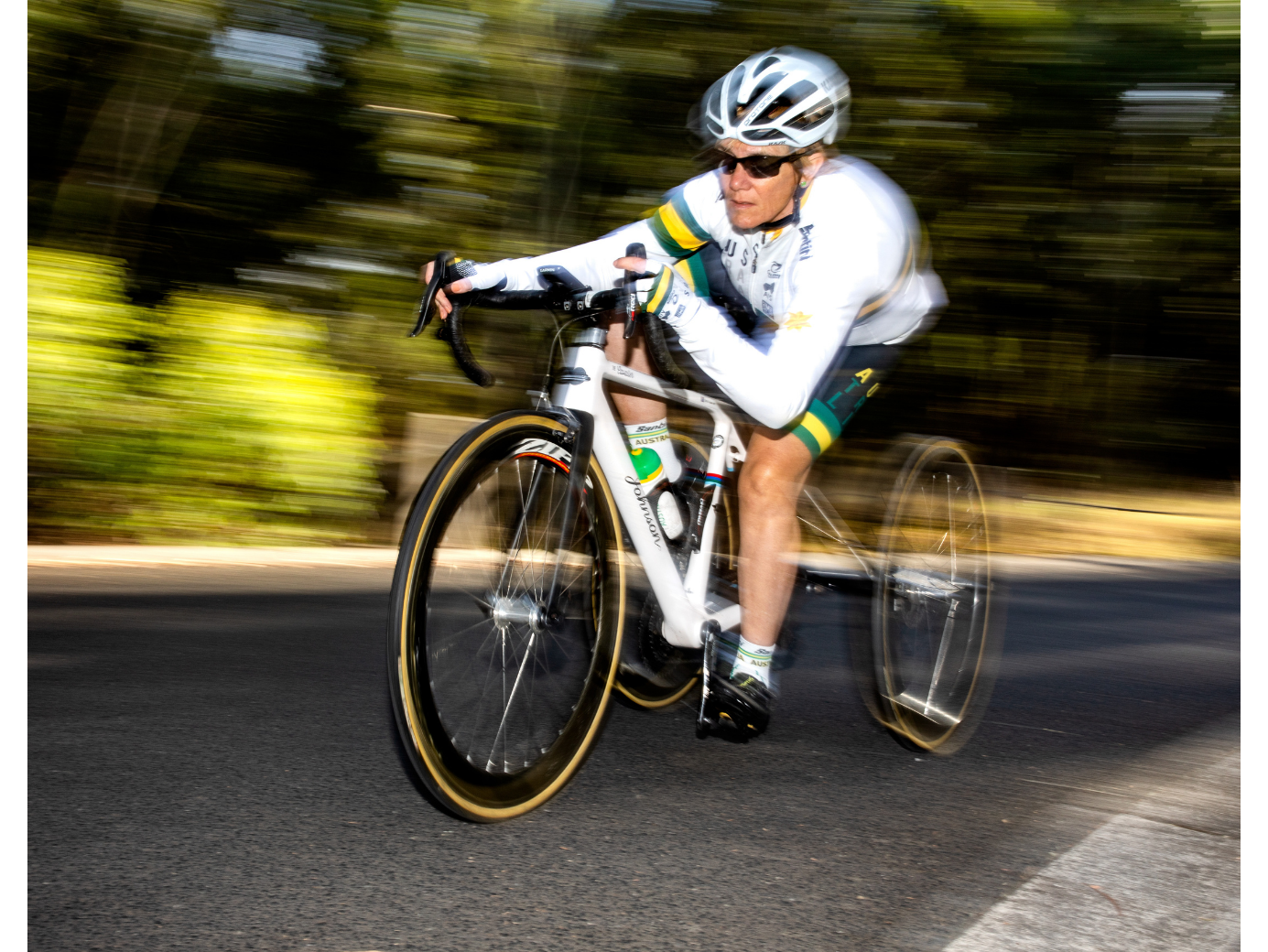 Carol in a white helmet and jersey riding a road bike at speed, with a blurred green background.