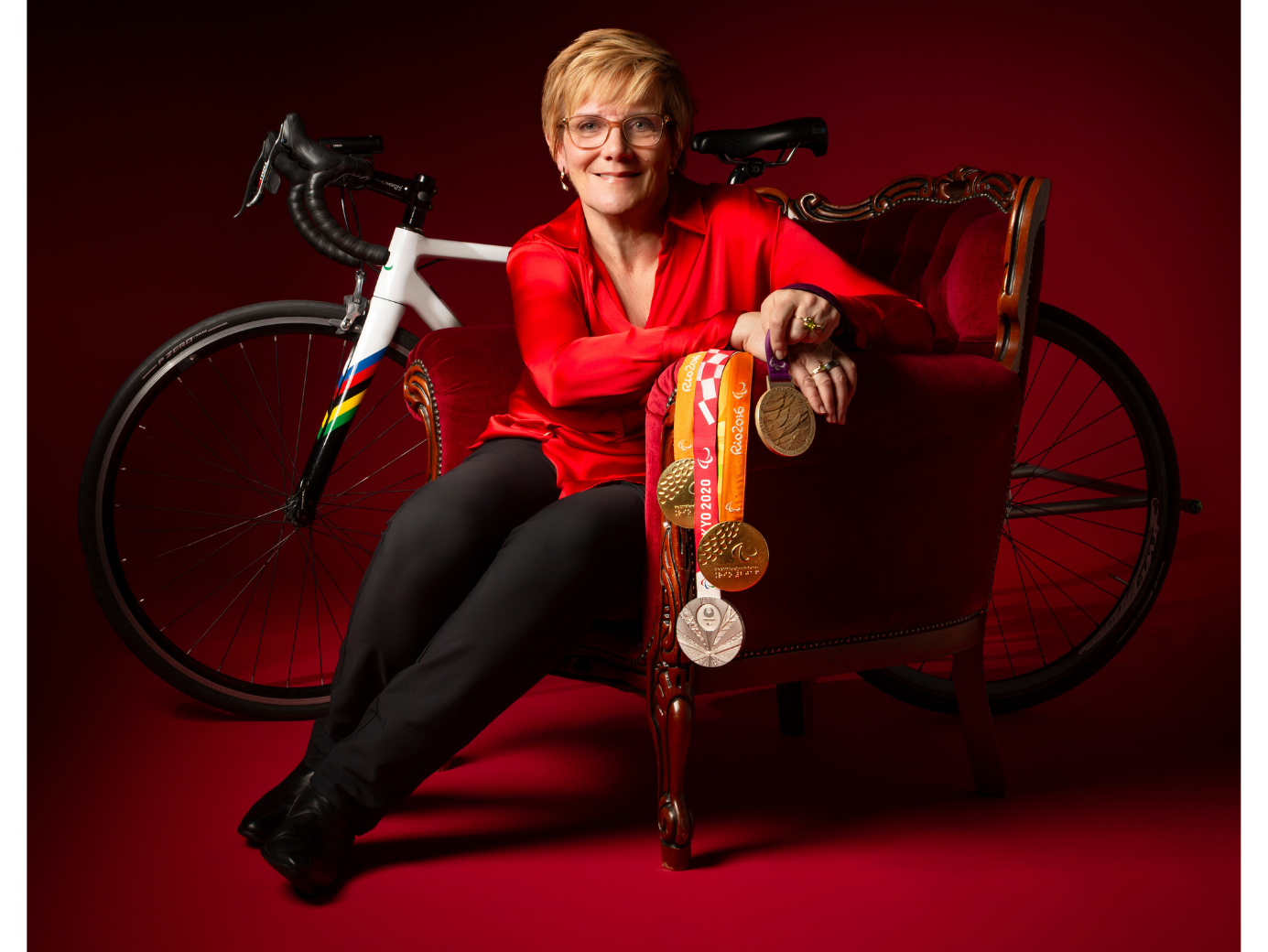 Carol in a red top and black pants sitting in a red armchair, holding medals, with a road bike behind her.