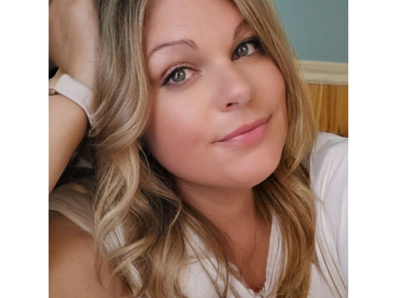 Michelle with long wavy blonde hair resting her head on her raised arm, wearing a white top.