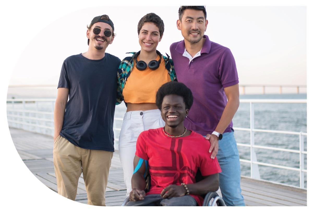 Four smiling young adults, one using a wheelchair, standing together on a waterfront boardwalk.