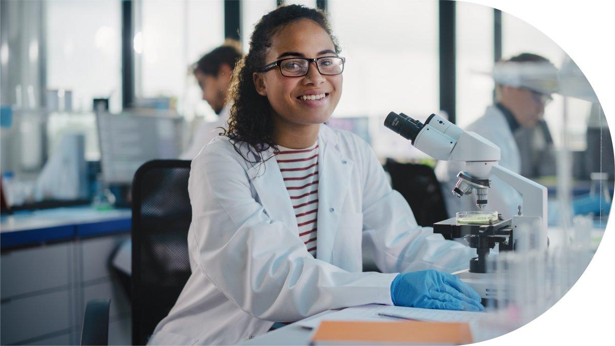 A smiling researcher in glasses and a lab coat, seated at a microscope in a laboratory.