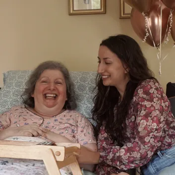 Stephanie, in a floral blouse, smiling at her mother, Carm, who is lying in bed in a pink pajama top with pink balloons in the background.