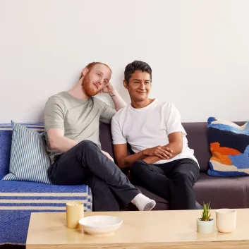 Marc, in a light green t-shirt and dark pants, sitting on a couch next to Richard, who is wearing a white t-shirt and dark pants.