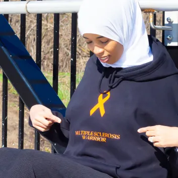 Najma sitting and looking down while pointing at black hoodie that reads 'Multiple Sclerosis Warrior.'