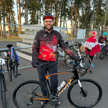 Brennan smiling while standing in front of his bike. He's wearing a helmet, glasses, black gloves, black pants, and a black and red jacket. A fence and bikes up against it are in the background.