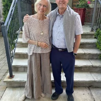 Jan and Tom, smiling in front of some steps outside. Jan is wearing a tan coloured shirt and skirt and Tom is wearing a light blue shirt, grey sweater around his shoulders, and blue pants..