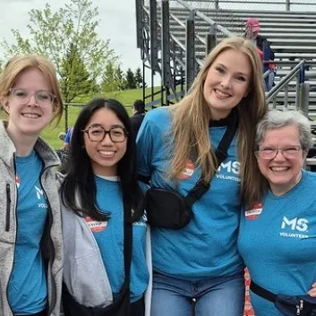 Four smiling women outdoors, three wearing blue MS charity shirts, at an outdoor event.