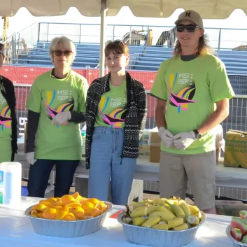 Four volunteers in green T-shirts standing behind a table with fruit at an outdoor event.